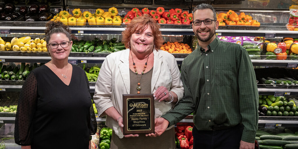 Two women and one man standing in grocery store produce section, holding award plaque.